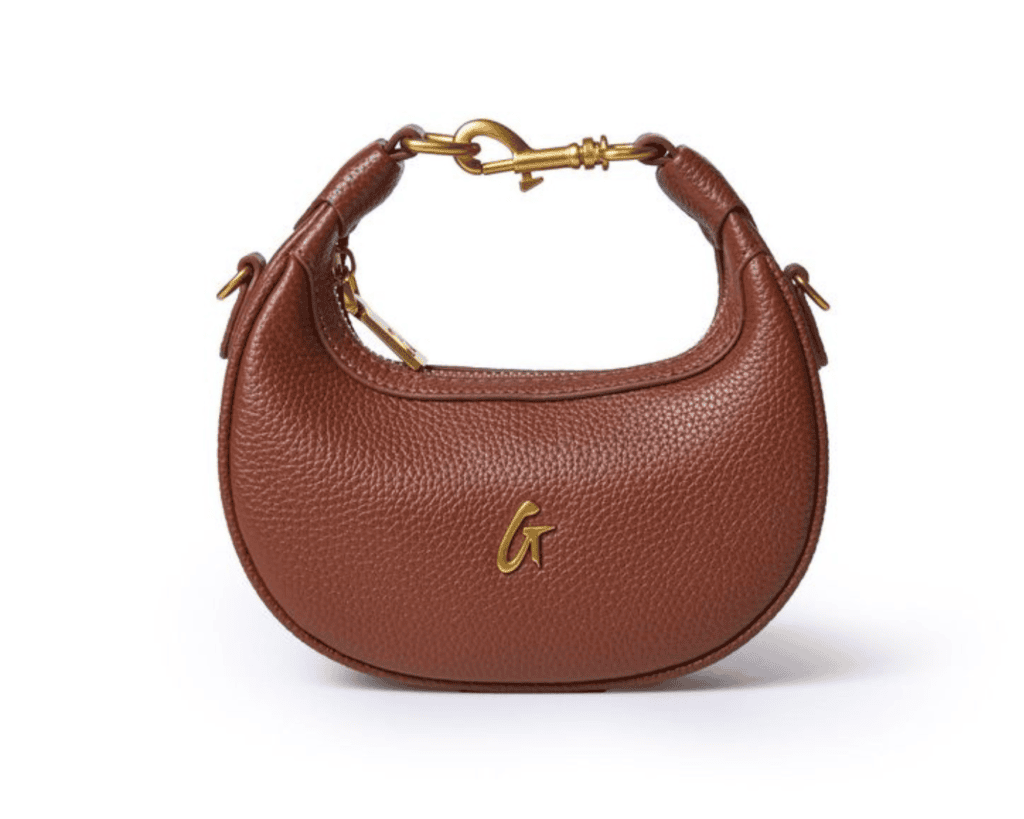 A studio front shot of a brown Glam-Aholic Lifestyle Nano Pebble Hobo bag featuring a curved crescent-shaped silhouette in textured pebbled vegan leather, a cursive gold-tone "G" script monogram logo centered on the front, and a unique top handle secured with a prominent gold swivel snap hook clasp.