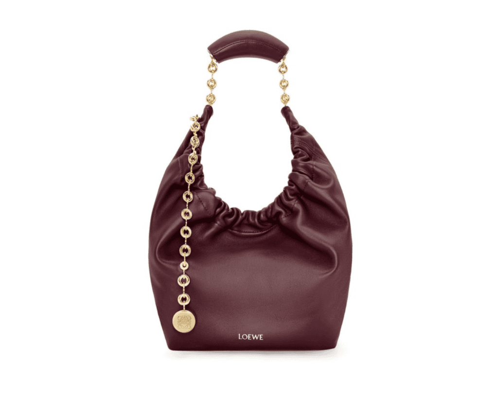 A studio front shot of a burgundy LOEWE Squeeze Small bag made of ultra-soft nappa lambskin leather, featuring its characteristic draped croissant silhouette, a ruched opening, a padded leather handle linked to a gold-tone donut chain strap, and an elongated decorative chain accent ending with an Anagram-engraved coin pendant.