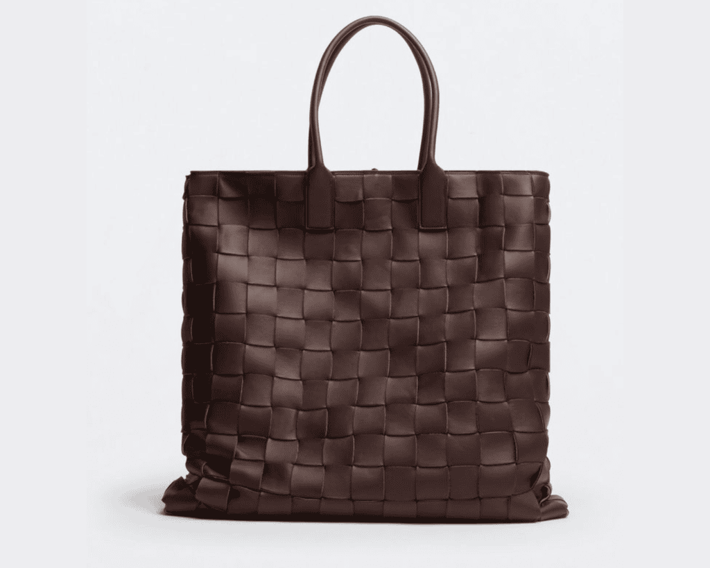 A studio front shot of a structured, square-shaped dark brown Bottega Veneta tote bag featuring an oversized, hand-woven maxi Intreccio leather pattern, complete with two integrated slim rolled leather top handles.
