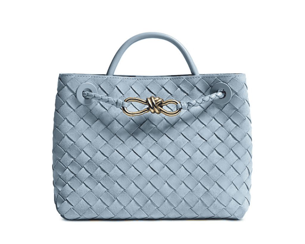 A studio front shot of a pale mineral blue Bottega Veneta Andiamo tote bag crafted in an all-over classic Intrecciato woven suede finish, featuring a structured top handle and a braided crossbody strap adorned with a signature sculptural gold-tone metal knot on the front.