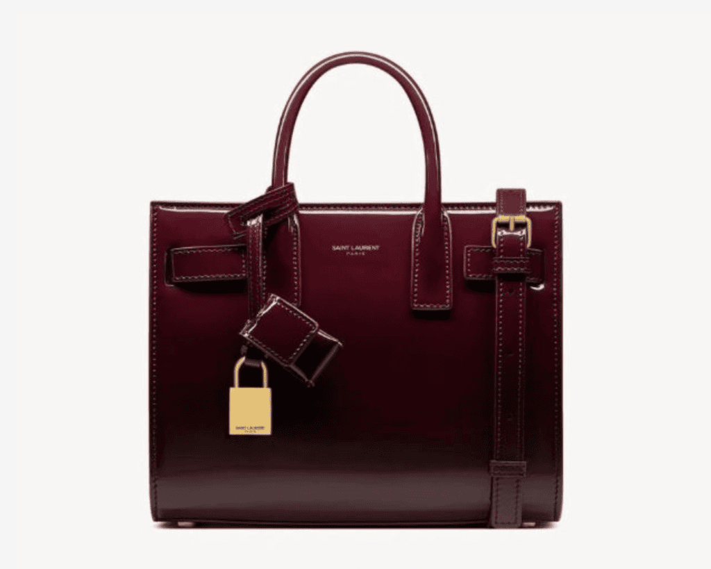A deep burgundy-red patent leather Saint Laurent Sac de Jour Nano satchel bag Mytheresa featuring structured tubular top handles Level Shoes, a flat leather shoulder strap Mytheresa, distinctive accordion-pleated side panels Saint Laurent, and an attached gold-tone padlock charm Saint Laurent.