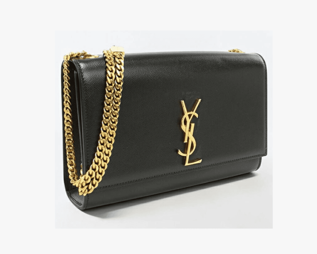 A side-angle studio shot of a black Saint Laurent Kate shoulder bag made of textured grain de poudre leather, featuring a prominent gold-tone interlocking YSL logo on the front flap and a chunky gold-toned gourmette chain strap resting on the top.